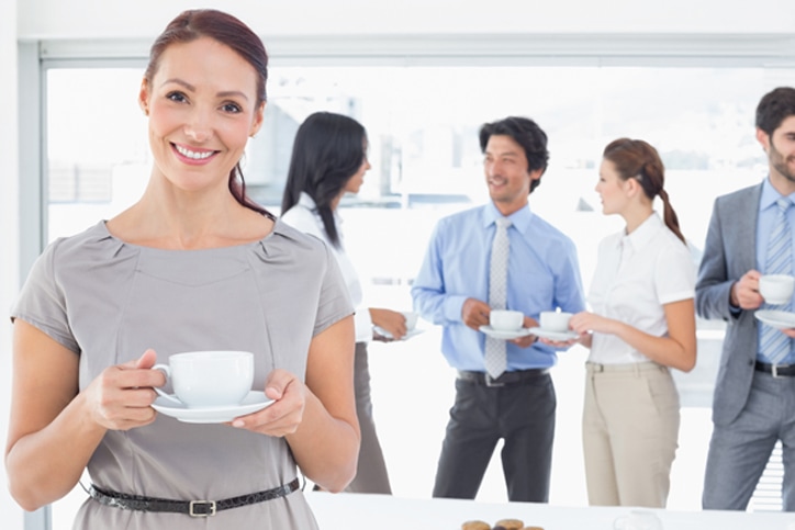 A smiling real estate agent holding a coffee with colleagues talking in the background.
